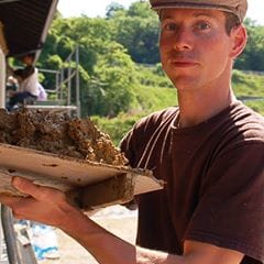 A natural building student in a cap holding a tray of wet clay at Daruma