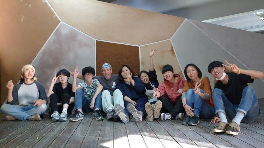 Natural building course group sitting on a wooden deck in front of a large earthen and clay plastered wall panel